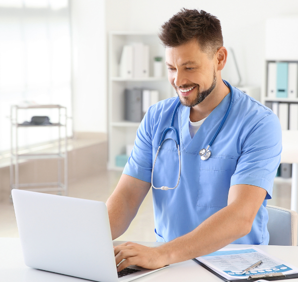 A male nurse practitioner sits at a computer and is smiling.